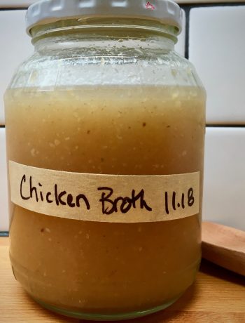 homemade chicken broth in a clear glass bottle with a lid labeled chicken broth on a wood counter with a wooden spoon