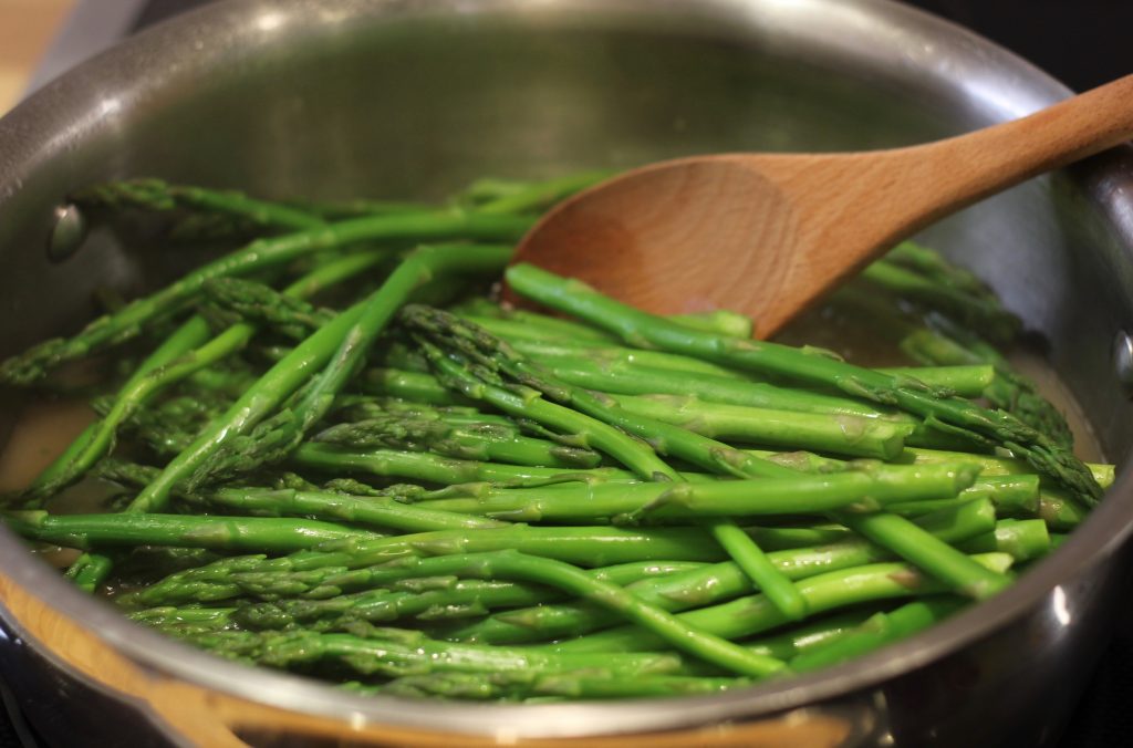asparagus simmering in chicken broth with wood spoon in stainless steel pan