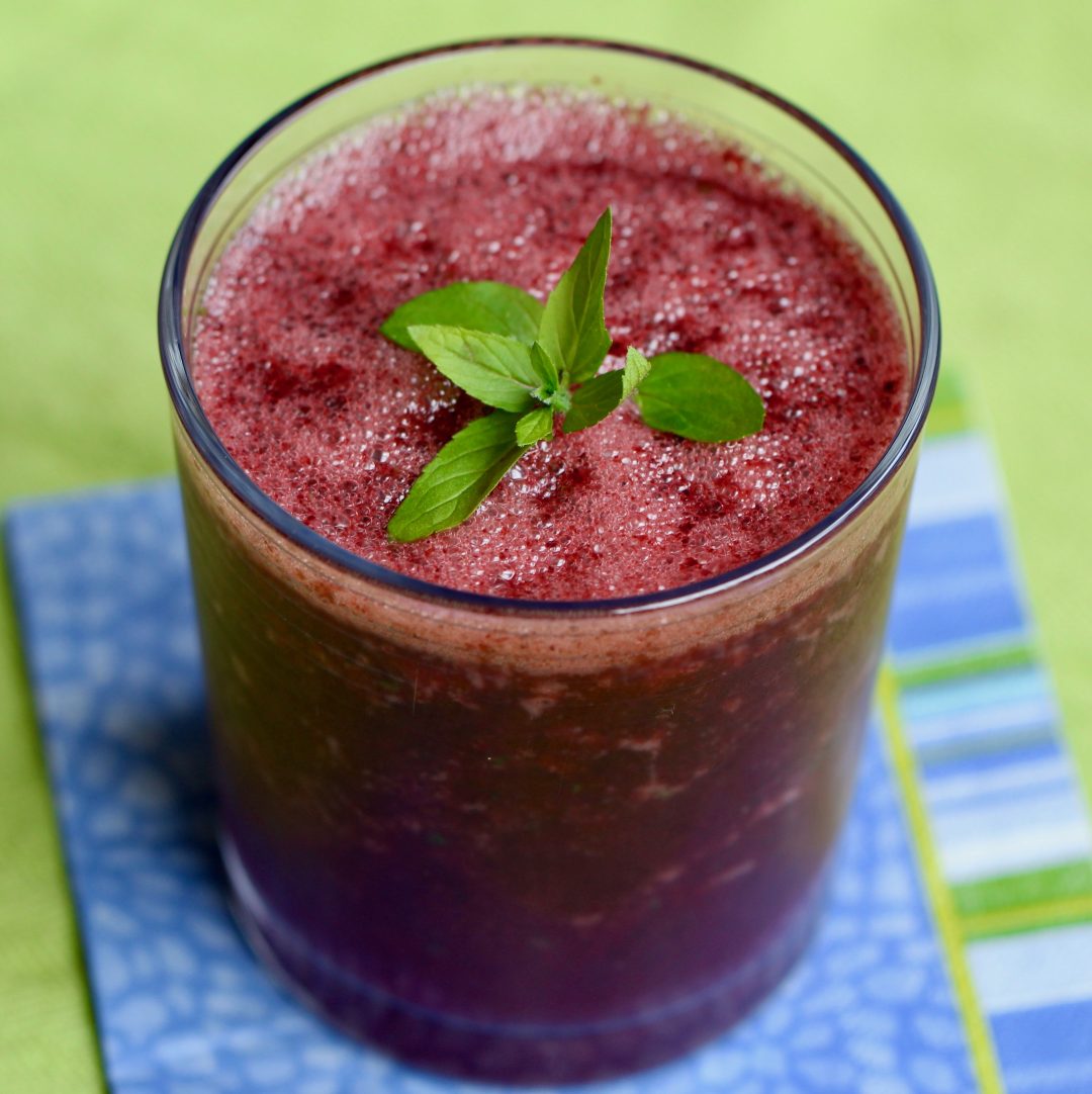 blueberry mint cooler in a glass with a sprig of mint on top on a blue napkin and green cloth