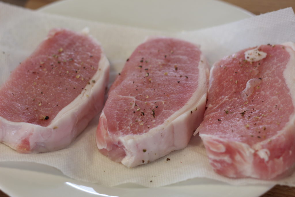 three raw pork chops sprinkled with pepper on a white ceramic plate