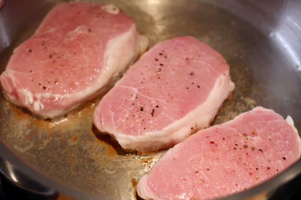 three raw pork chops cooking in a stainless steel pan