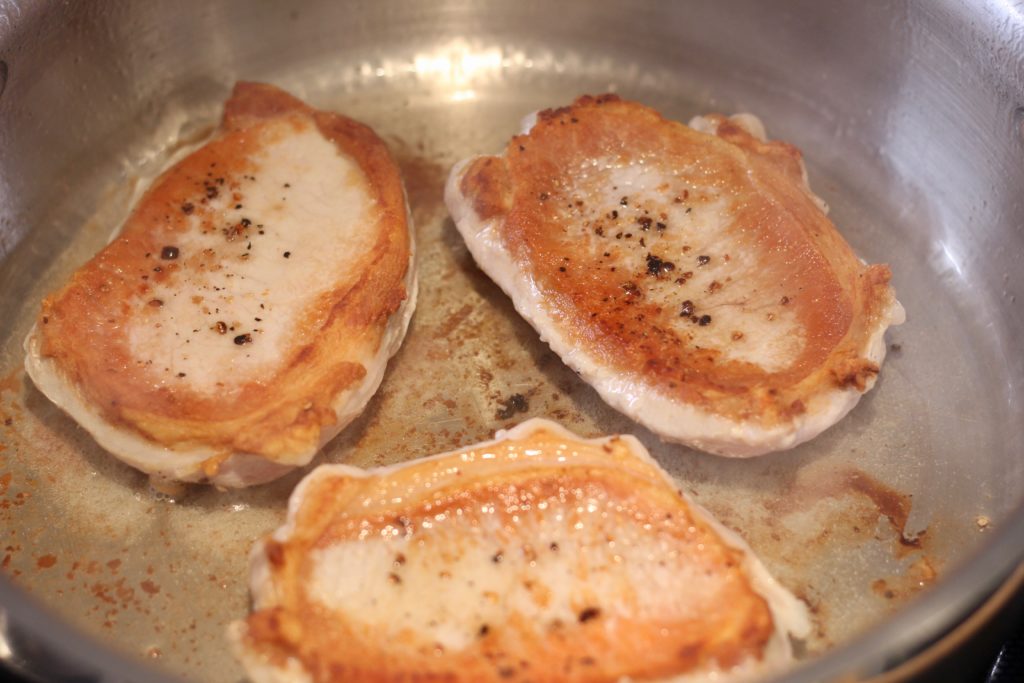 three pork chops cooking in a stainless steel pan
