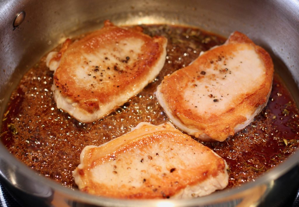 three browned pork chops cooking in bubbling apple cider vinegar glaze in a stainless steel pan