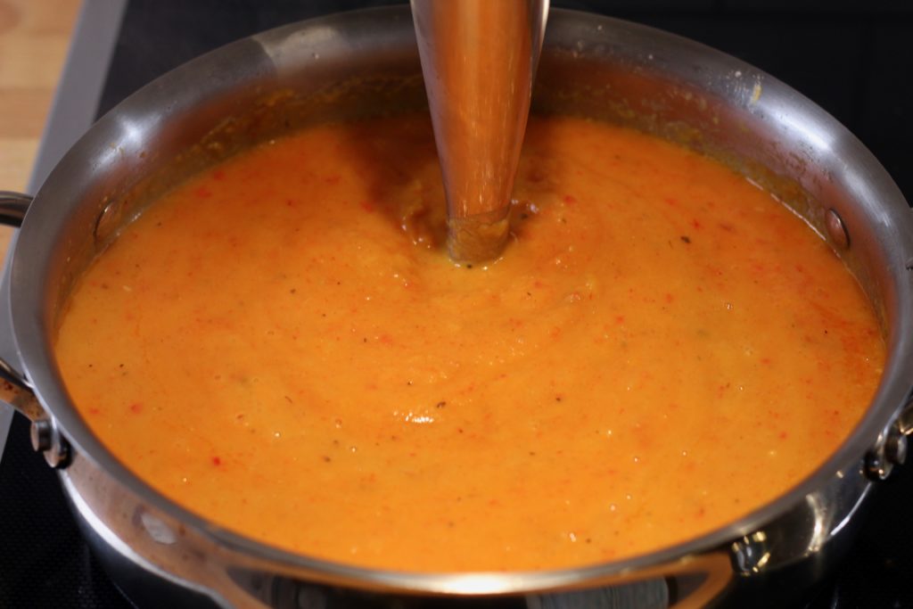 pumpkin soup with apples and red peppers being blended with an immersion blender in a stainless steel pan